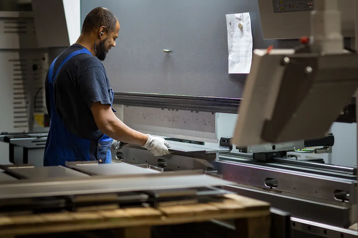 Man in een werkplaats bedient een industriële zetbank en buigt metalen platen, terwijl hij handschoenen en werkkleding draagt.