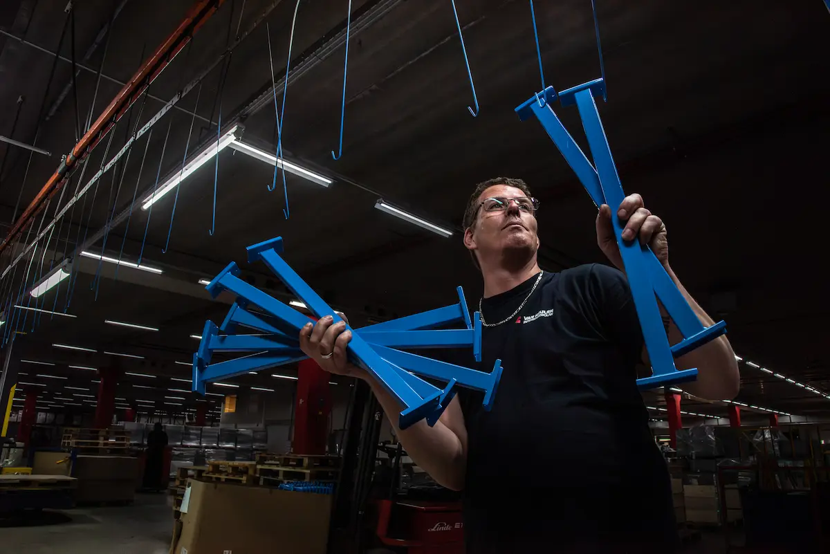 Medewerker in een fabriekshal houdt meerdere blauwe metalen onderdelen vast, terwijl boven hem haken aan een transportsysteem hangen.