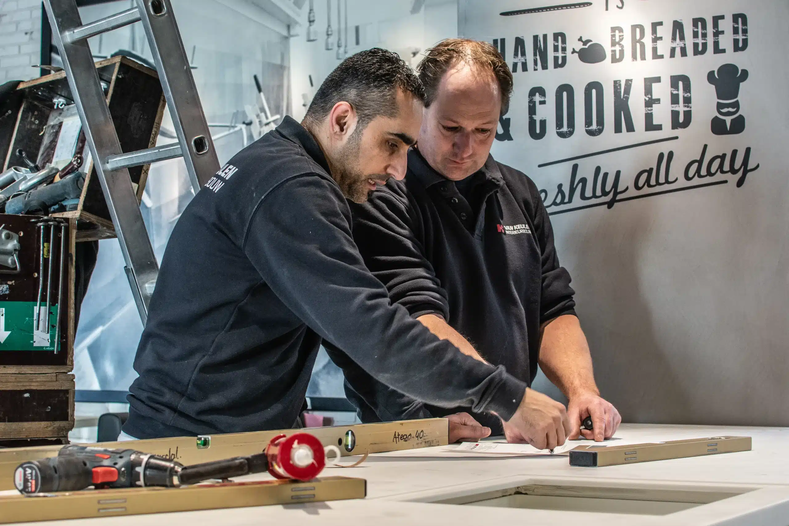 Twee mannen werken samen aan een werkbank, waarbij ze nauwkeurig meten en tekenen op een houten plank, in een werkplaatsomgeving.