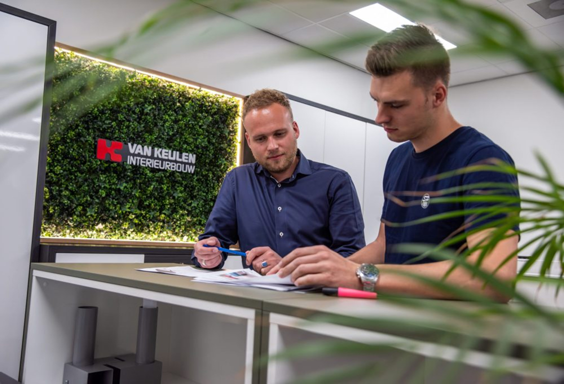 Twee medewerkers van Van Keulen Interieurbouw bespreken ontwerpen en documentatie in een moderne kantoorruimte met groene wanddecoratie.